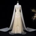 A formal gown displayed on a mannequin against a black background, featuring a cream tulle dress with extensive gold and silver beadwork and a matching sheer cream cape with a long train.