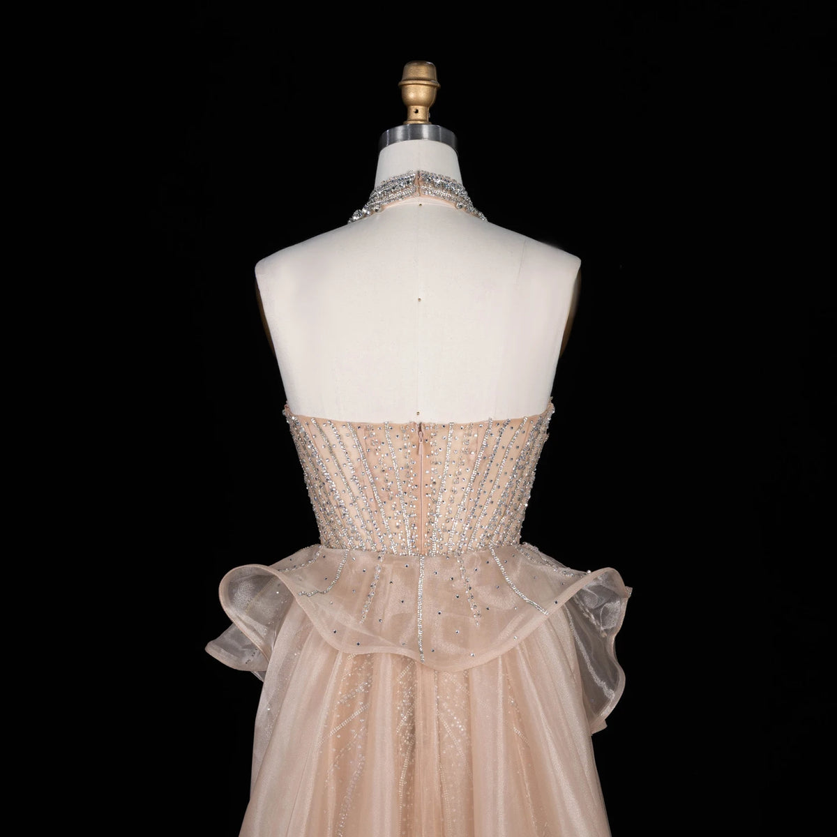 A formal gown displayed on a white dress form against a black background. The dress has a strapless bodice covered in silver rhinestone embellishments, a sheer ruffled waist, and a layered tulle skirt in a pale blush pink color.