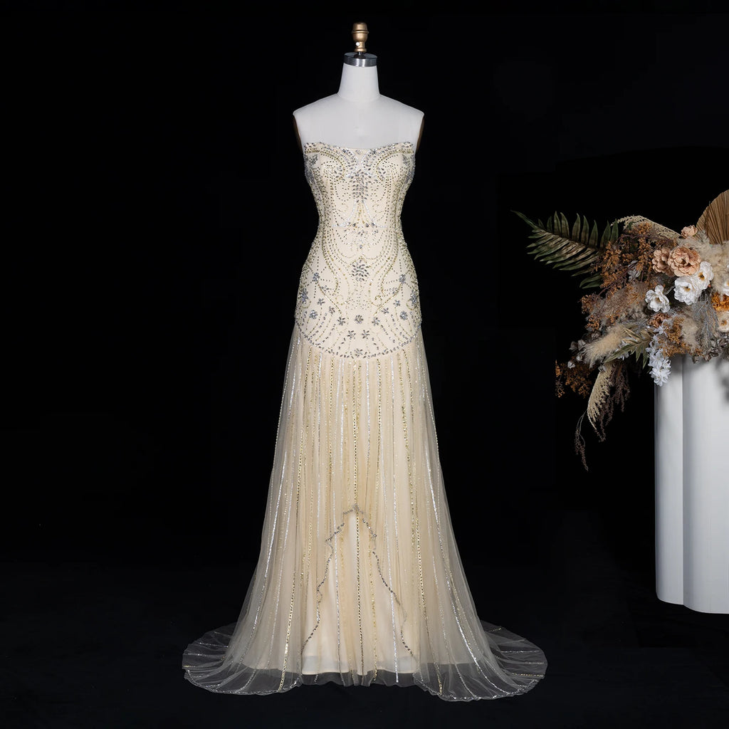 A strapless formal gown displayed on a mannequin against a black background, with decorative dried flowers at the right edge. The dress is champagne beige with intricate silver and clear beadwork and embellishments across the bodice and skirt.