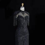 A black beaded evening gown displayed on a dress form mannequin against a solid black background. The dress features a high neckline with sheer mesh panels, intricate beadwork, fringe details on the sleeves, and a fitted silhouette.