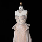 A strapless formal gown displayed on a cream mannequin against a black background. The dress is embellished with dense silver beadwork and sequins, featuring a sweetheart neckline, a fitted corset-style bodice, a sheer ruffled overlay at the waist, and a flowing skirt.
