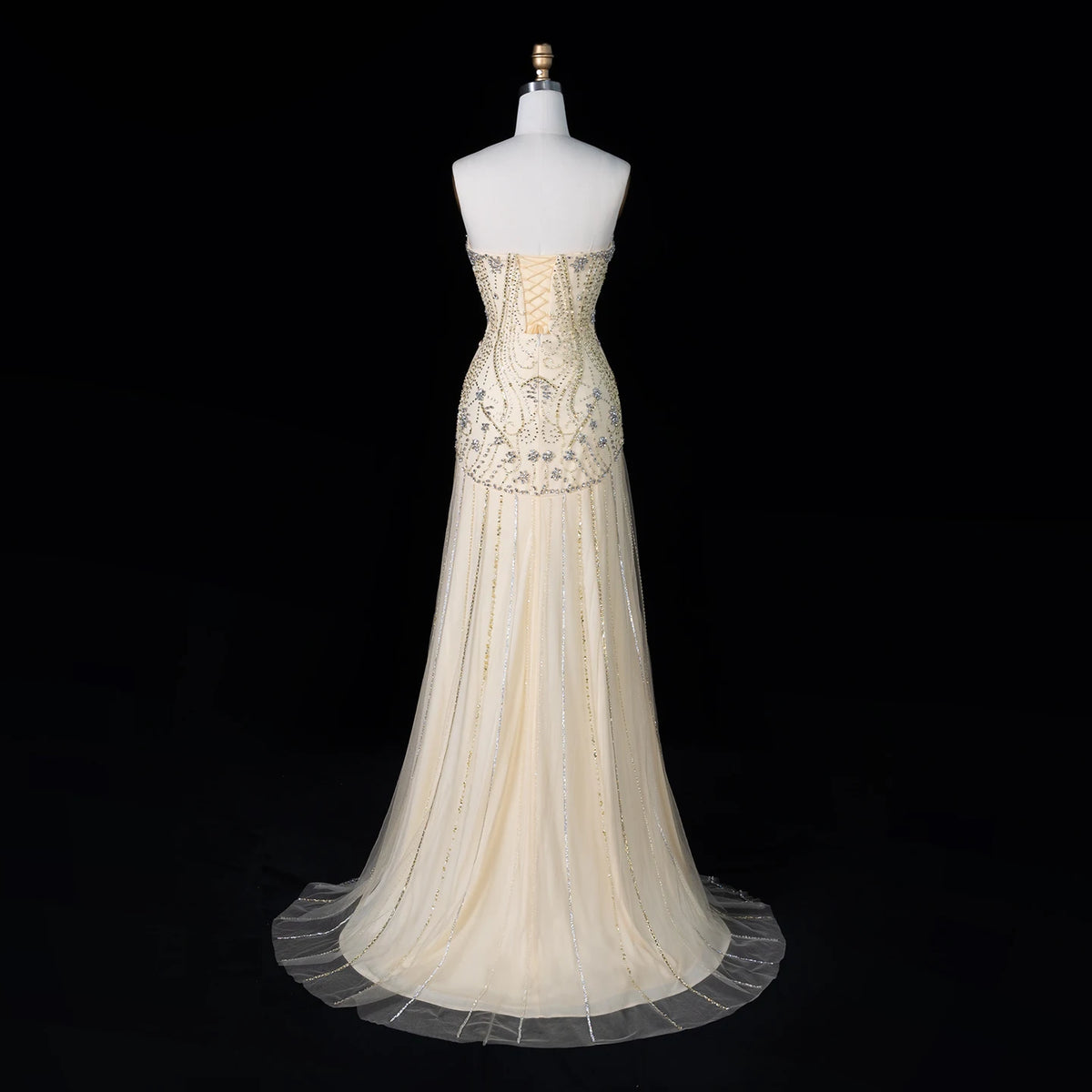 A strapless formal gown displayed on a white dress form against a black background. The dress is a champagne or ivory color with intricate gold beading and embroidery concentrated on the bodice and waist, and a sheer tulle skirt with a long train.